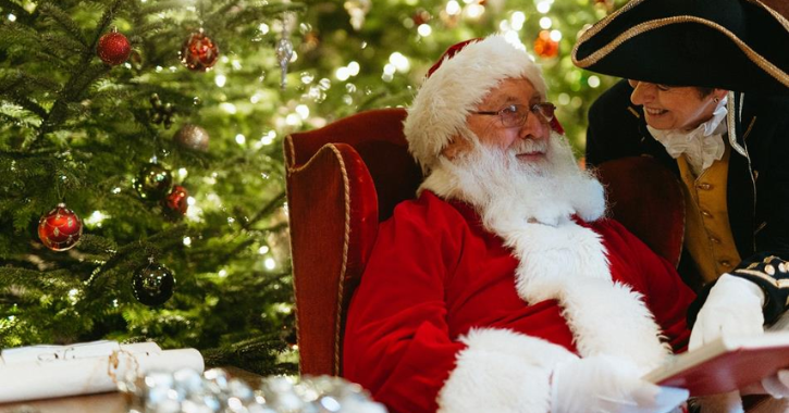 Father Christmas sat in chair with a footman smiling next to him, with twinkling Christmas lights in background.